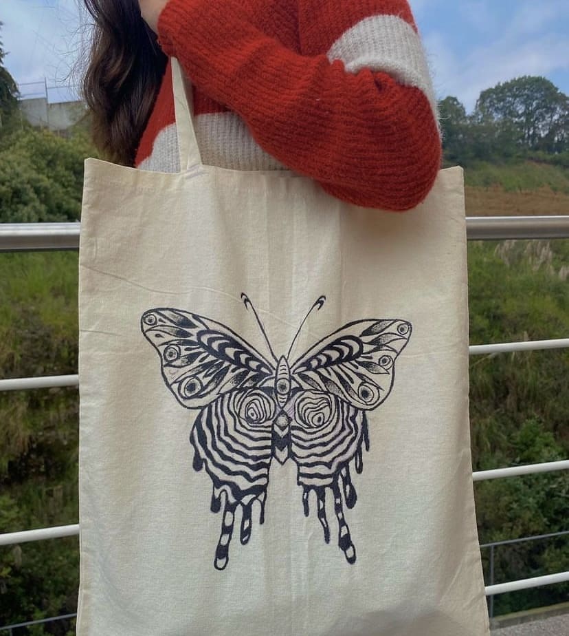 Fotografía de tote bag con diseño de mariposa pintada a mano con pintura negra, tomada en el exterior con un fondo de naturaleza, en que la persona portadora utiliza un suéter café con blanco.